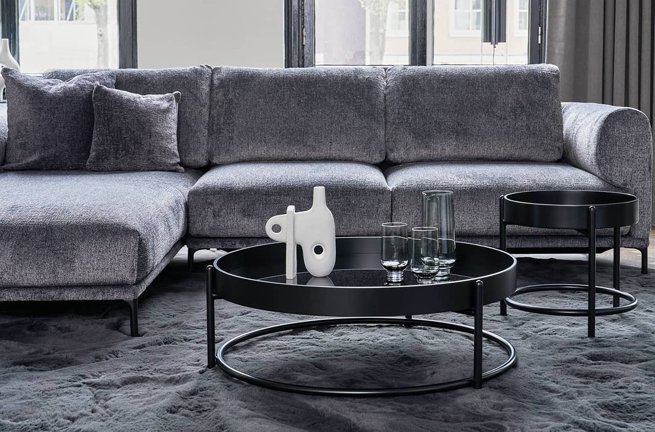 Schwarzer runder Couchtisch mit flacher Tischplatte und filigranem Metallgestell in moderner Wohnumgebung mit grauem Sofa und dunklem Teppich.